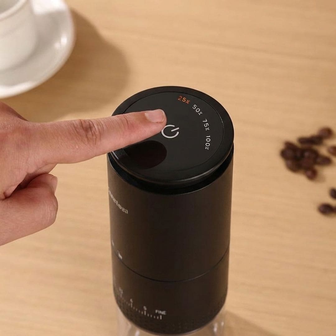 A close-up of a finger pressing the touch power button on top of the Gustosa Electric Coffee Grinder, with the LED display showing battery level indicators at 25%, 50%, 75%, and 100%, next to scattered coffee beans on a wooden surface.