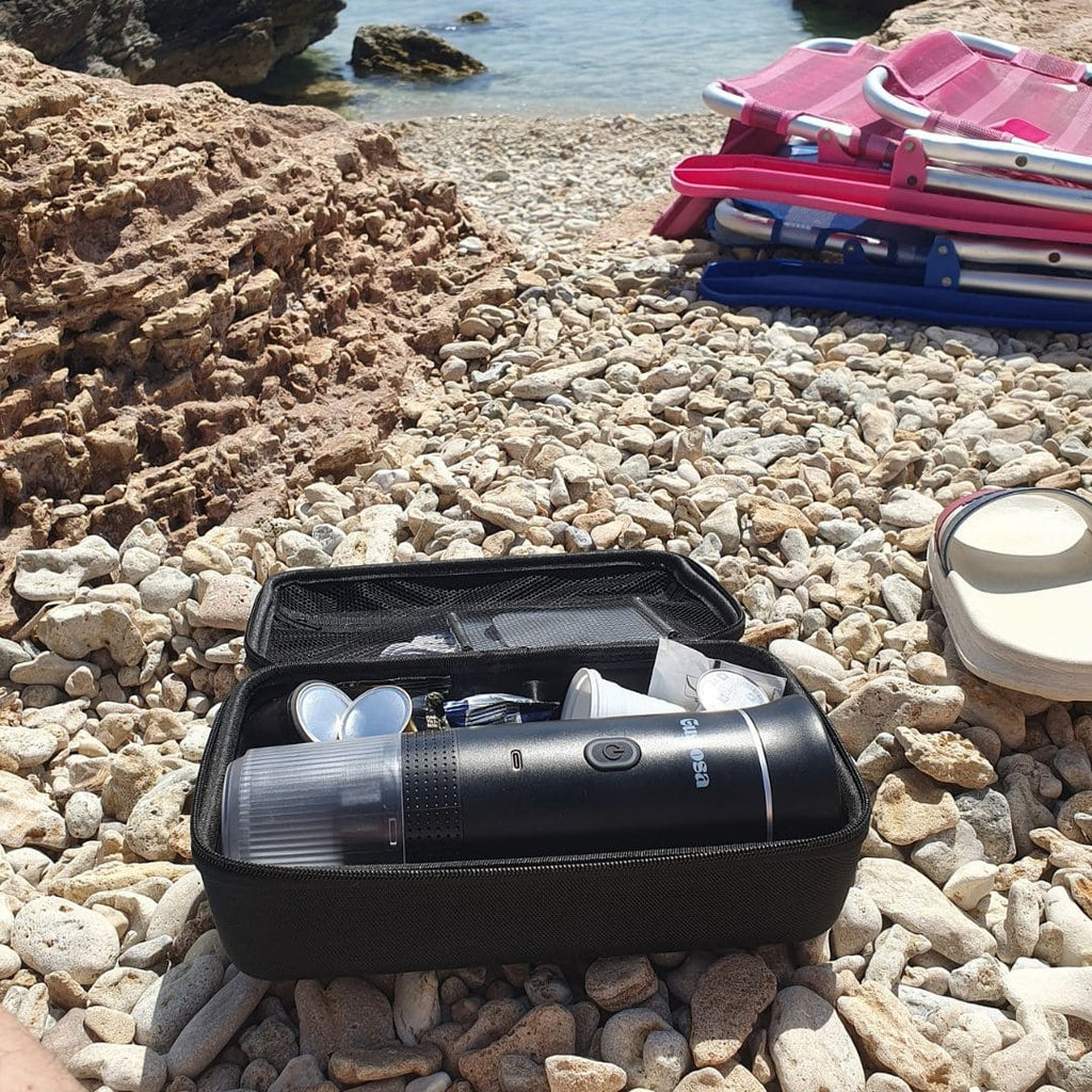 The Gustosa Bag open on a rocky Mediterranean beach, showing MyGustosa portable espresso maker neatly stored inside alongside cups and accessories, demonstrating how organized and travel-ready the bag keeps your entire coffee kit.