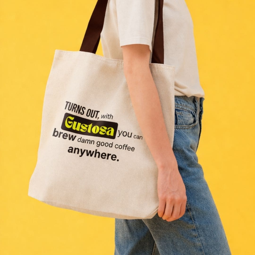 A person carrying the Gustosa canvas shopper tote bag with the brand slogan "Turns out, with Gustosa you can brew damn good coffee anywhere" printed on the front, shown against a bright yellow background.