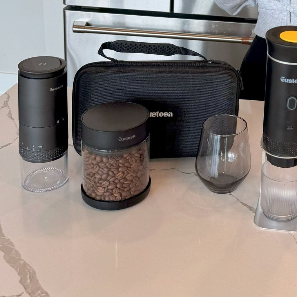 The complete Gustosa coffee ecosystem displayed on a marble kitchen counter, including the Gustosa Electric Grinder, Vacuum Container filled with coffee beans, Gustosa Bag, a glass cup, and MyGustosa portable espresso maker.