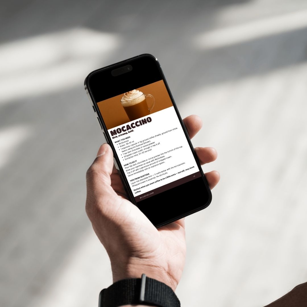 A hand holding a smartphone displaying the Mocaccino recipe page from the Gustosa Special Recipes eBook, showing the recipe photo, ingredients and step-by-step instructions, demonstrating how easy it is to access the guide on any device.