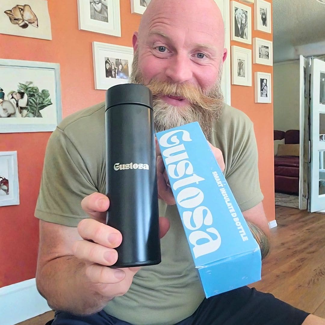 A smiling bearded man holding up the Gustosa Smart Bottle in matte black alongside its branded blue packaging box, capturing the unboxing moment of the Gustosa Smart Insulated Bottle.