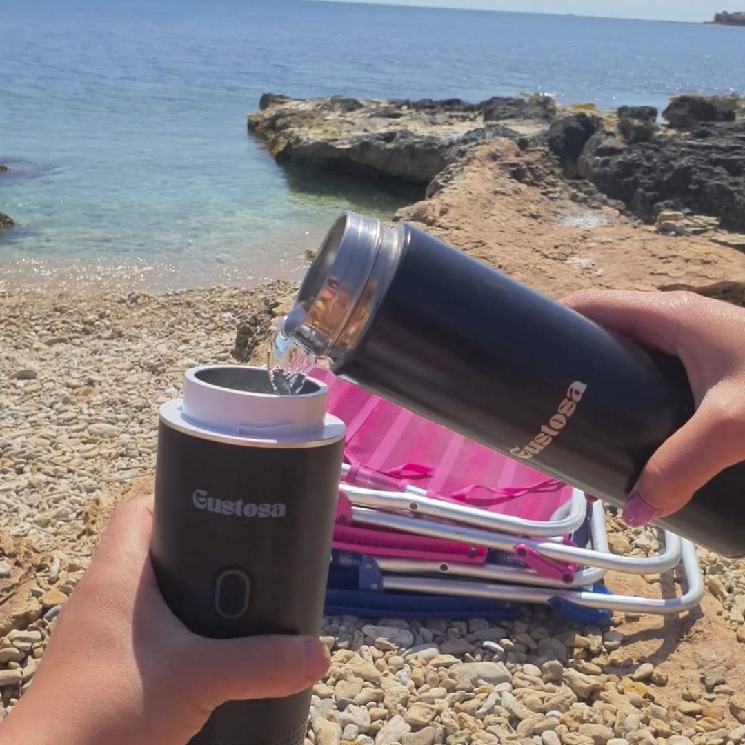 A person pouring water from the Gustosa Smart Bottle into MyGustosa portable espresso maker on a rocky beach, showing how the two products work together to brew fresh coffee anywhere outdoors.