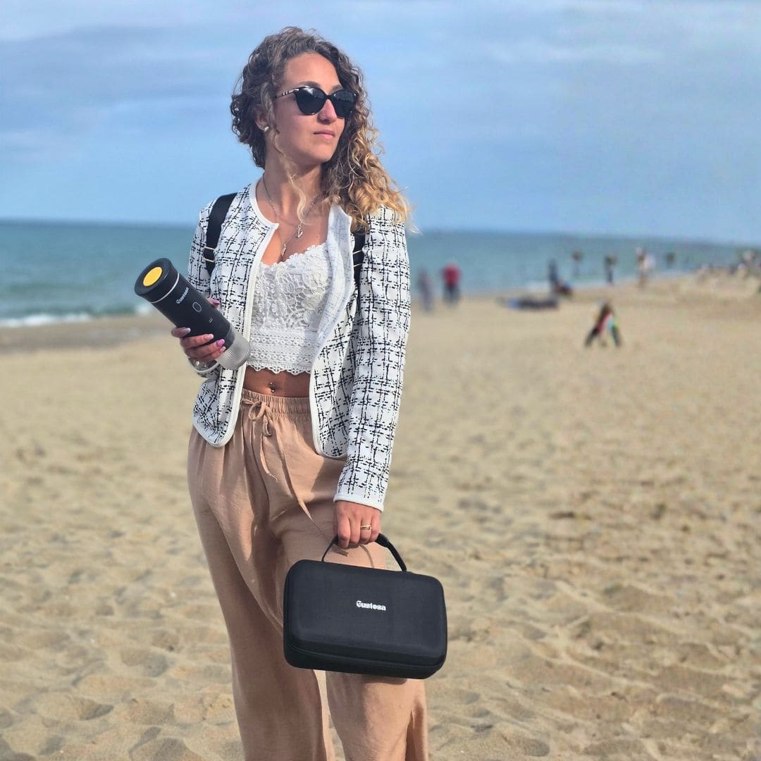 A stylish woman standing on a sandy beach holding MyGustosa portable espresso maker in one hand and the Gustosa Bag in the other, showing how easy it is to bring your complete coffee setup anywhere.