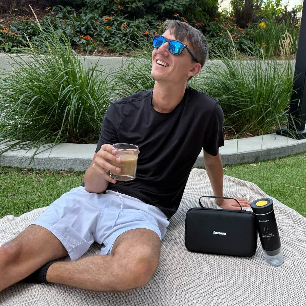 Smiling man relaxing outdoors with iced coffee, Gustosa portable coffee maker, and travel bag beside him on a picnic blanket