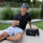 Smiling man relaxing outdoors with iced coffee, Gustosa portable coffee maker, and travel bag beside him on a picnic blanket